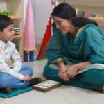 Teacher sitting on the floor with a child during a hands-on activity at Krish Elite preschool in ECIL